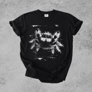 Puede incluir: Una camiseta negra de manga corta con un estampado gr&aacute;fico blanco de una ara&ntilde;a saltarina peluda en el pecho. La ilustraci&oacute;n de la ara&ntilde;a tiene un aspecto desgastado y texturizado con salpicaduras de tinta visibles alrededor.