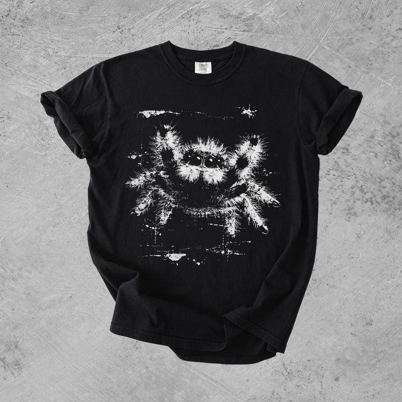Puede incluir: Una camiseta negra de manga corta con un estampado gr&aacute;fico blanco de una ara&ntilde;a saltarina peluda en el pecho. La ilustraci&oacute;n de la ara&ntilde;a tiene un aspecto desgastado y texturizado con salpicaduras de tinta visibles alrededor.