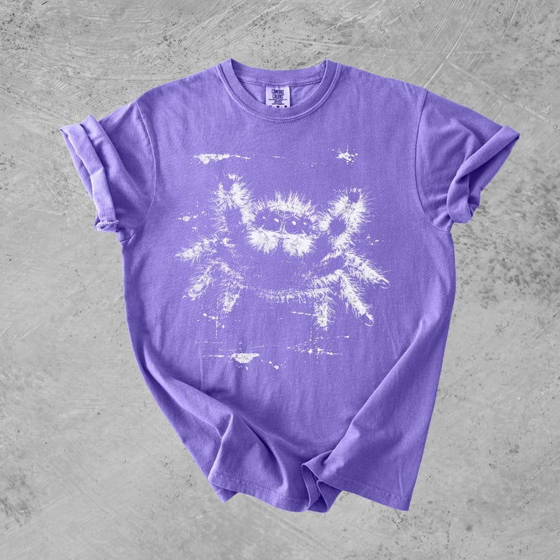 Puede incluir: Una camiseta morada de manga corta con un estampado gr&aacute;fico blanco de estilo desgastado de una ara&ntilde;a saltarina en la parte delantera. La prenda tiene cuello redondo y una etiqueta en el interior del cuello que dice Comfort Colors.