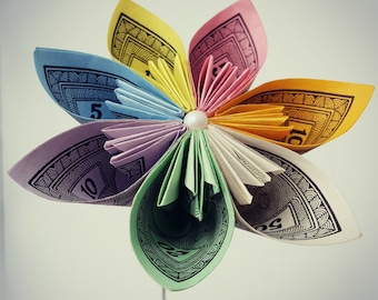 May include: A colorful paper flower made from folded playing cards. The flower has ten petals, each with a different color and design. The center of the flower is decorated with a small white pearl.