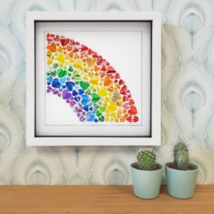 Op de afbeelding: Een ingelijst regenboogkunstwerk gemaakt van kleurrijke hartvormige papieren stukjes. Het kunstwerk wordt tentoongesteld op een muur met een groen-wit gestreept behang. Twee kleine cactusplanten in potten staan op de voorgrond.