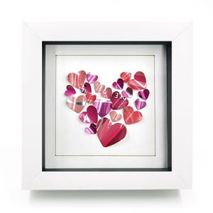 Può includere: Un'opera d'arte incorniciata con un cuore composto da ritagli a forma di cuore rosa e rossi. I cuori sono disposti su uno sfondo bianco, all'interno di una cornice bianca con un bordo interno nero. Un pezzo decorativo.