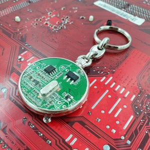 May include: A green circuit board keychain with a silver ring. The circuit board has a variety of electronic components, including resistors, capacitors, and integrated circuits. The keychain is on a red background.