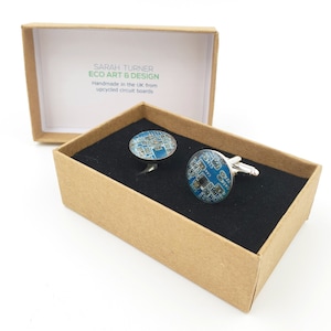 Op de afbeelding: Een paar manchetknopen in een bruine doos met een zwarte binnenkant. De manchetknopen zijn gemaakt van gerecyclede printplaten en hebben een blauw en zilver ontwerp. De doos heeft een label met de tekst "SARAH TURNER ECO ART & DESIGN Handgemaakt in het Verenigd Koninkrijk van gerecyclede printplaten".