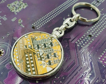 May include: A silver key chain with a round, yellow and black patterned charm. The charm resembles a circuit board.