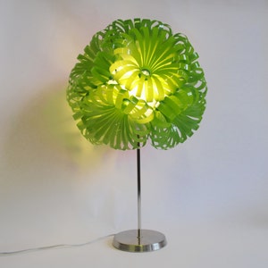 May include: A green table lamp with a unique, flower-shaped shade made of plastic strips. The lamp has a silver metal base and a cord.