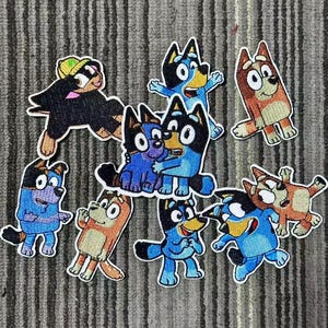 May include: A collection of embroidered patches featuring cartoon dog characters in various poses. The patches are in shades of blue, brown, black, and yellow, with white outlines. The patches are likely for clothing or accessories.