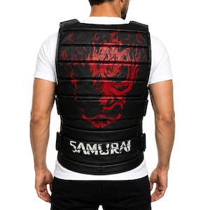 May include: A black padded vest featuring horizontal stitching and a large red graphic of a stylized mask on the back. The word SAMURAI is printed in white distressed lettering across the lower section of the vest, which includes side adjustment straps.