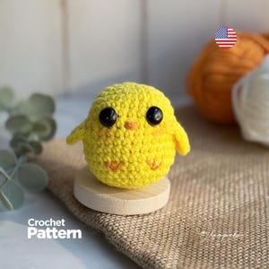 May include: A small, round, bright yellow crocheted chick amigurumi featuring two large black safety eyes, a tiny orange stitched beak, and two small orange stitched feet. The chick is mounted on a circular light-coloured wooden base.