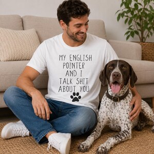 Op de afbeelding: Een wit t-shirt met korte mouwen en zwarte tekst die luidt MY ENGLISH POINTER AND I TALK SHIT ABOUT YOU, vergezeld van een zwarte pootafdruk. Het shirt wordt gedragen met een blauwe spijkerbroek en witte sneakers.