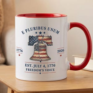 Pode incluir: Uma caneca de cerâmica branca com interior e alça vermelhos. A frente apresenta um gráfico do Liberty Bell com um padrão de bandeira americana, o texto E PLURIBUS UNUM, 1776, 2026, EST. JULY 4, 1776 e FREEDOM'S VOICE, com estrelas azuis.