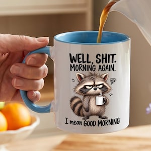 Könnte beinhalten: Ein weißer Keramikbecher mit hellblauem Innenraum und Henkel, verziert mit einer Grafik eines mürrischen Waschbären, der eine kleine Kaffeetasse hält. Der Text lautet: WELL, SHIT. MORNING AGAIN. I mean GOOD MORNING. Dunkle Flüssigkeit wird eingegossen.