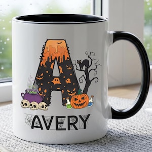 Puede incluir: Una taza de cerámica blanca con asa y borde interior negros. El frente presenta una letra A grande decorada con goteos naranjas, fantasmas y calabazas, junto a un gato negro, un caldero morado y una calabaza. El nombre AVERY está impreso debajo.