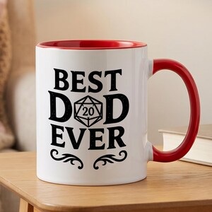 May include: A white ceramic mug with a red interior and handle. The front features black text that reads BEST DAD EVER, with a 20-sided die graphic replacing the letter A in DAD. The design includes decorative flourishes at the bottom.
