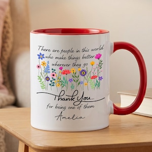 Può includere: Una tazza in ceramica bianca con interno e manico rosso brillante. L'esterno presenta un'illustrazione colorata di fiori di campo e il testo stampato: There are people in this world who make things better wherever they go. Thank You... for being one of them. Amelia.