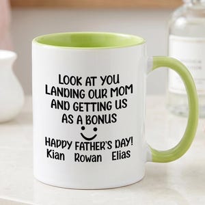 Puede incluir: Taza de cerámica blanca con interior y asa verdes. La taza presenta texto negro que dice "LOOK AT YOU LANDING OUR MOM AND GETTING US AS A BONUS" con una carita sonriente y "HAPPY FATHER'S DAY!" seguido de tres nombres.