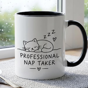 Puede incluir: Una taza de cerámica blanca con interior y asa negros, que presenta un dibujo lineal negro de un gato durmiendo, el texto "PROFESSIONAL NAP TAKER", tres símbolos "z" y dos pequeños iconos de corazón impresos en la parte frontal.