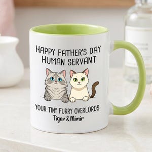 May include: A white ceramic mug with a light green interior and handle. The front features a graphic of two cats with the text: Happy Father's Day Human Servant, Your Tiny Furry Overlords, Tiger & Mimir, printed in black lettering.