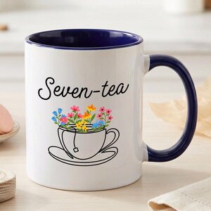 Può includere: Una tazza in ceramica bianca con interno e manico blu scuro. La parte anteriore presenta il testo nero "Seven-tea" sopra un'illustrazione a linee di una tazza da tè riempita con fiori colorati rosa, gialli e blu e una bustina di tè.
