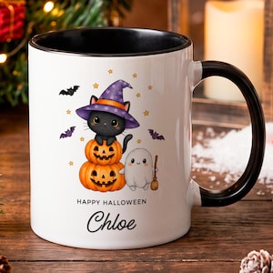 Puede incluir: Una taza de cerámica blanca con asa e interior negros, que presenta un gráfico de un gato negro con sombrero de bruja morado sobre dos calabazas naranjas junto a un pequeño fantasma blanco con una escoba, con el texto Happy Halloween Chloe.
