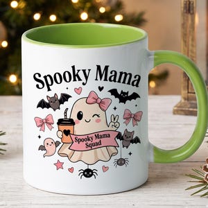 May include: A white ceramic mug with a lime green interior and handle. The front features a cartoon ghost with a pink bow, holding a coffee cup, surrounded by bats, spiders, and pink bows. The text reads Spooky Mama and Spooky Mama Squad.