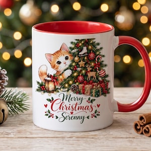 Puede incluir: Una taza de cerámica blanca con interior y asa rojos, que presenta un gráfico de un gatito atigrado naranja asomándose detrás de un árbol de Navidad decorado con regalos, una linterna y el texto Merry Christmas Serenny en letra roja y verde.
