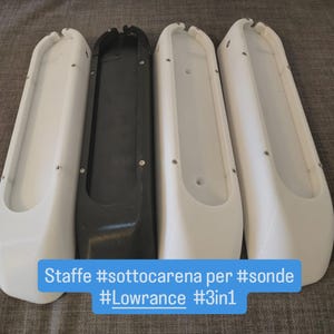 Può includere: Quattro supporti per trasduttori per barca. Tre sono bianchi e uno nero. I supporti sono lunghi e stretti con estremità arrotondate e diversi fori per viti. Testo su sfondo blu: "Staffe #sottocarena per #sonde #Lowrance #3in1".