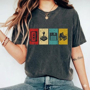 May include: Dark gray t-shirt featuring a retro design with a cassette tape, joystick, floppy disk, and roller skate in red, yellow, blue, and yellow rectangles. The shirt has a vintage, distressed look.