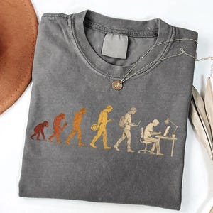Peut inclure: Un t-shirt &agrave; col rond gris anthracite avec un imprim&eacute; graphique dans des tons orange et jaune. Le motif illustre l'&eacute;volution d'un primate vers un humain moderne assis &agrave; un bureau, travaillant sur un ordinateur avec une lampe de bureau.