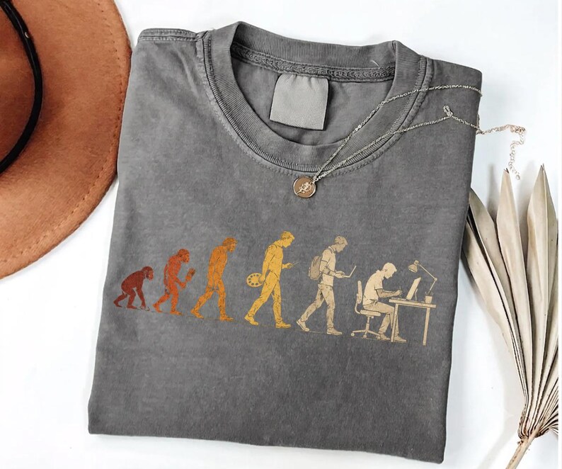 Peut inclure: Un t-shirt &agrave; col rond gris anthracite avec un imprim&eacute; graphique dans des tons orange et jaune. Le motif illustre l'&eacute;volution d'un primate vers un humain moderne assis &agrave; un bureau, travaillant sur un ordinateur avec une lampe de bureau.