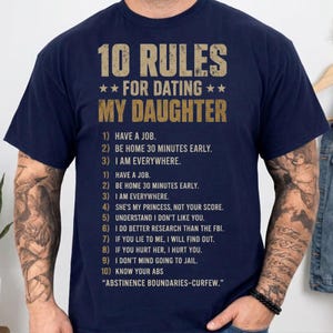 May include: A navy blue short-sleeved t-shirt featuring the text 10 RULES FOR DATING MY DAUGHTER in a distressed tan font, followed by a numbered list of ten rules and the phrase ABSTINENCE BOUNDARIES-CURFEW printed in a matching tan color.