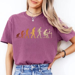 Peut inclure: Un t-shirt &agrave; manches courtes de couleur prune avec une impression graphique dor&eacute;e illustrant l'&eacute;volution de l'humanit&eacute;, passant d'un primate &agrave; un individu moderne assis &agrave; un bureau avec un ordinateur et une lampe.