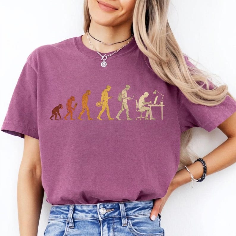 Peut inclure: Un t-shirt &agrave; manches courtes de couleur prune avec une impression graphique dor&eacute;e illustrant l'&eacute;volution de l'humanit&eacute;, passant d'un primate &agrave; un individu moderne assis &agrave; un bureau avec un ordinateur et une lampe.
