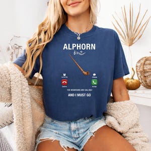 May include: Navy blue t-shirt with the word "ALPHORN" in white, a musical note, and a brown alphorn illustration. Below, it reads "THE MOUNTAINS ARE CALLING! AND I MUST GO" with phone icons.