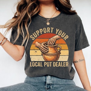 May include: A charcoal gray short-sleeve t-shirt featuring a graphic print of hands shaping a clay pot. The design includes a sunset-colored background with horizontal stripes and the text "SUPPORT YOUR LOCAL POT DEALER" in a bold, arched font.