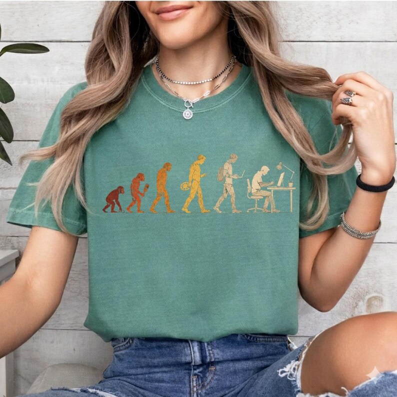 Peut inclure: Un t-shirt &agrave; col rond vert sauge avec une impression graphique de l'&eacute;volution humaine, partant d'un primate pour aboutir &agrave; une personne assise &agrave; un bureau avec un ordinateur. Le motif est rendu dans des tons de jaune et d'orange sur le devant.