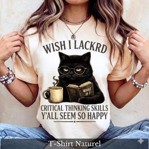 Puede incluir: Una camiseta de color crema con el gráfico de un gato negro con gafas redondas, sosteniendo una taza de café y un libro abierto. El texto dice: WISH I LACKRD CRITICAL THINKING SKILLS Y'ALL SEEM SO HAPPY. La portada del libro tiene el texto: WHAT IF 2.