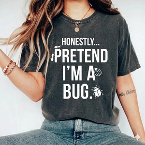 Könnte beinhalten: Dunkelgraues T-Shirt mit dem weißen Text "EHRLICH... TU SO, ALS WÄRE ICH EIN KÄFER." Das Design zeigt Illustrationen einer Ameise, eines Marienkäfers und eines Käfers. Ein lässiger Unisex-Stil.