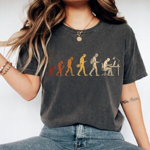 Peut inclure: Un t-shirt gris anthracite &agrave; manches courtes avec une impression graphique repr&eacute;sentant l'&eacute;volution humaine. Le motif montre une progression d'un primate vers un humain moderne assis &agrave; un bureau avec un ordinateur, dans des tons orange, jaune et blanc.