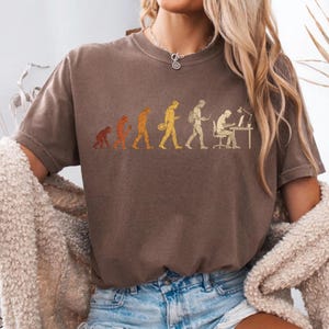 Peut inclure: Un t-shirt marron &agrave; manches courtes avec une impression graphique repr&eacute;sentant l'&eacute;volution humaine, partant d'un primate et progressant par &eacute;tapes jusqu'&agrave; une personne assise &agrave; un bureau avec un ordinateur, le tout dans des tons jaune et orange.