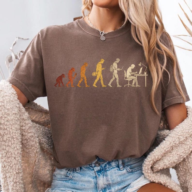 Peut inclure: Un t-shirt marron &agrave; manches courtes avec une impression graphique repr&eacute;sentant l'&eacute;volution humaine, partant d'un primate et progressant par &eacute;tapes jusqu'&agrave; une personne assise &agrave; un bureau avec un ordinateur, le tout dans des tons jaune et orange.