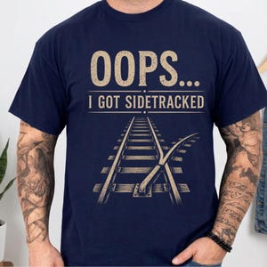 Peut inclure: Un t-shirt bleu marine à manches courtes avec une impression graphique de couleur beige clair. Le motif comprend le texte OOPS... I GOT SIDETRACKED au-dessus d'une illustration de voie ferrée qui se divise en deux directions sur le devant.