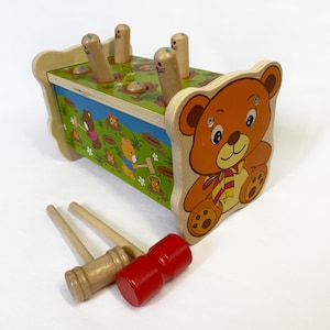 May include: Wooden toy featuring a bear design. The toy has a green top with wooden pegs and a colorful illustration of bears. Includes three wooden mallets, one red and two natural wood.