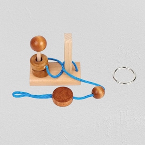 Può includere: Un puzzle in legno con una base marrone chiaro, un palo verticale e una palla di legno. Due anelli di legno e una palla di legno sono collegati da una corda blu. È incluso anche un anello d'argento.