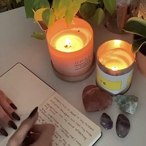 Peut inclure: Deux bougies allumées, une dans un pot en verre orange et l'autre dans un récipient en métal, sont posées sur une table. Un carnet avec du texte manuscrit, des cristaux et une pierre en forme de cœur sont à proximité.