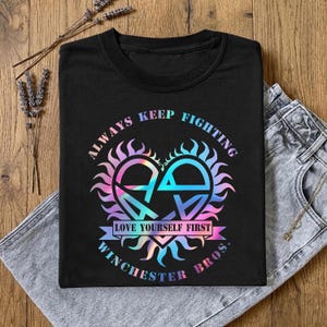 Może przedstawiać: Czarny t-shirt z kolorowym wzorem graficznym. Wzór przedstawia emblemat w kształcie serca z napisami "ALWAYS KEEP FIGHTING", "LOVE YOURSELF FIRST" i "WINCHESTER BROS."