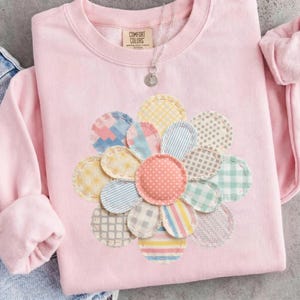 May include: A light pink sweatshirt with a large, appliqued flower design. The flower petals are made from various patterned fabrics in pastel colors. The sweatshirt has a crew neck and long sleeves. The tag reads "Comfort Colors". A silver necklace with a pendant is visible.