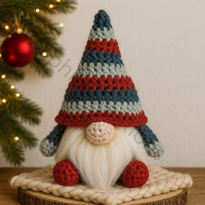 Op de afbeelding: Een handgemaakte kabouterpop met een gestreepte rood, blauw en grijs gehaakte hoed, witte baard en een beige neus. De kabouter heeft rode gehaakte voeten en armen. Hij zit op een houten schijf en een beige gehaakte mat. Kerstdecoratie.