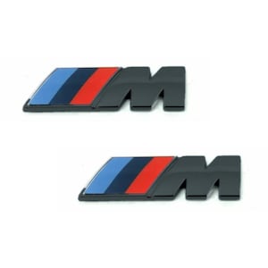 May include: Two black car emblems featuring the letter "M" and a rectangular design with blue, dark blue, and red stripes. The emblems are on a white background.