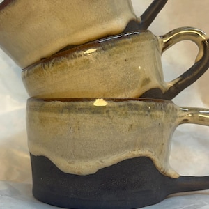 Könnte beinhalten: Ein Stapel aus drei Keramikbechern mit zweifarbigem Design. Der untere Teil jedes Bechers ist in dunklem, mattem Anthrazit gehalten, während der obere Bereich mit einer glänzenden, cremefarbenen Glasur versehen ist, die über die Basis läuft.